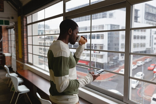 Man drinking coffee and using smartphone while looking out office window, copy space