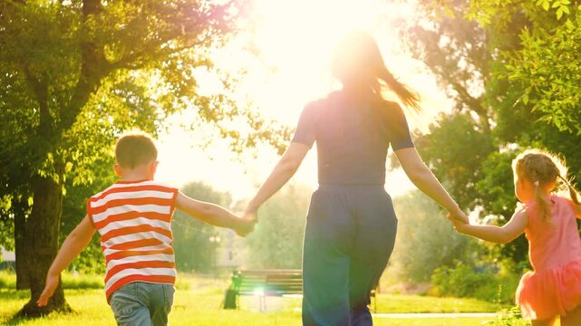 Silhouette of mother with hands interlocked with children leading across park lawn. Mother and children run across park with hands interlocked in bond of shared joy. Mother leads children through park