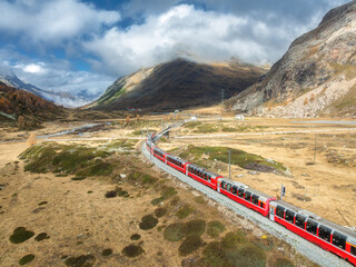 Aerial view of modern red train, alpine mountains, orange hills and trees, sky with clouds at sunny...