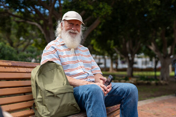 Elderly man with a long white beard and mustache, wearing a beige cap, striped short-sleeve sweater, and blue jeans, sitting on a wooden bench in a park, smiling while holding a smartphone