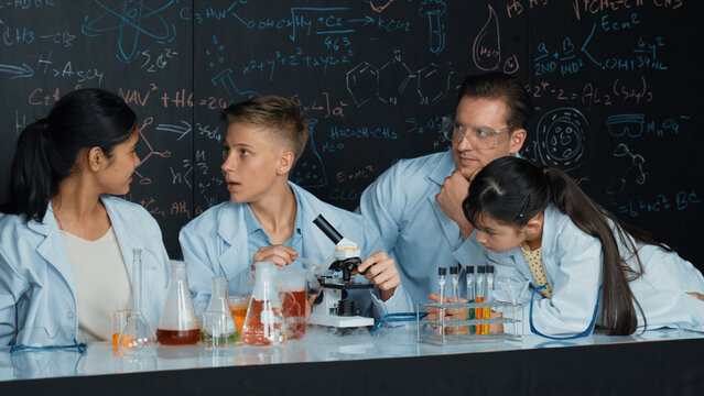 Multicultural student looking under microscope while cute girl inspect sample. Teacher looking after highschool children doing experiment at blackboard with science theory in STEM class. Edification.