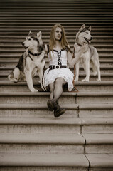 Naklejka premium Young woman with long blonde hair sits on steps with two siberian huskies. Dressed in white dress, she gazes ahead. Sepia tone creates nostalgic, urban atmosphere