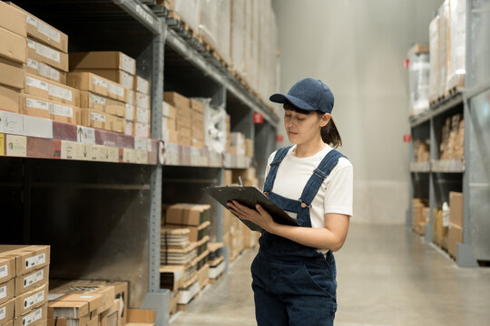 Warehouse worker wearing a hat checking quality by clipboard on tall shelves in warehouse storage. Auditor woman or staff work looking up stock taking inventory in cargo store. Logistic business work. - Powered by Adobe