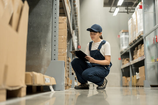Warehouse worker wearing a hat checking quality by clipboard on tall shelves in warehouse storage. Auditor woman or staff work looking up stock taking inventory in cargo store. Logistic business work.