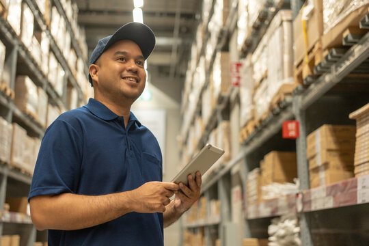 Warehouse worker wearing a hat and blue shirt hands holding tablet check stock on tall shelves in warehouse storage. Asian auditor or staff work looking up stocktaking inventory in warehouse store.
