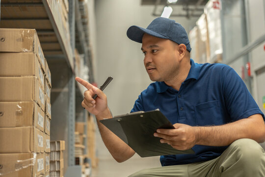 Warehouse worker wearing a hat and black shirt checking quality by clipboard on tall shelves in warehouse storage. Asian auditor or staff work looking up stock taking inventory in warehouse store.