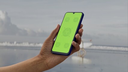 GREEN SCREEN CHROMA KEY A person interacts with a smartphone on a beach with a green screen, ideal for app mockups or digital interface overlays. Peaceful beach setting