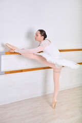 Young female ballerina with dark hair stretches leg on barre in dance studio. Wearing white tutu and practice attire. Bright, minimalistic room enhances focus on technique