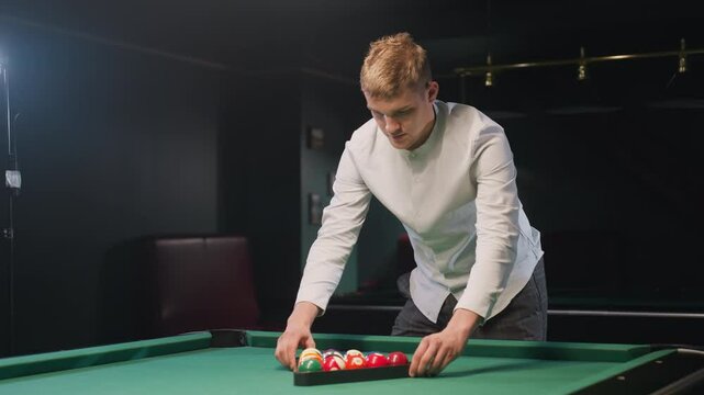 Billiard trainee in white shirt and gray trousers arranges colorful billiard balls in triangle formation on vibrant green pool table under warm ambient lighting with serious facial expression