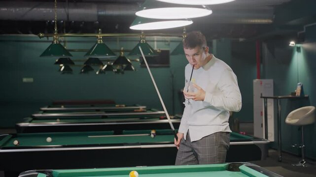 Close-up of a player in white shirt with calm expression, holding cue stick and glass of lemon drink while pacing around pool hall. Overhead lights cast a stylish ambiance over green pool tables