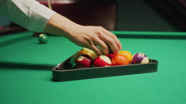 Person arranges colorful billiard balls inside triangle rack on green pool table, ensuring proper alignment before break shot. captures precision and focus in pool hall setting before game begins