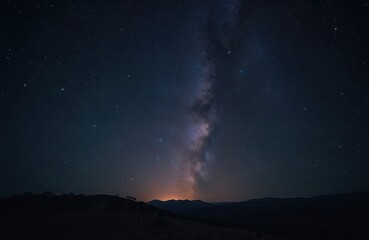 Night sky with bright Milky Way band and stars over dark mountain silhouette. Space cosmic background. Universe astro landscape wallpaper. Travel explore serene world. Starry night.