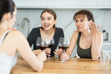 Three joyful ladies in their middle years chatting while having wine in goblets in the kitchen