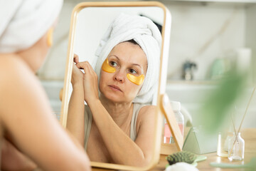 Relaxed woman in her middle age using under eye patches for skincare sitting at the mirror in home environment