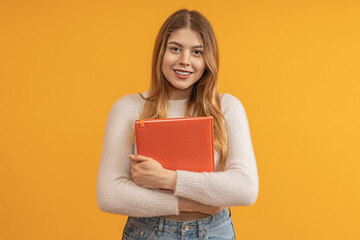 A young woman with long hair holding an orange folder, smiling, against a yellow background