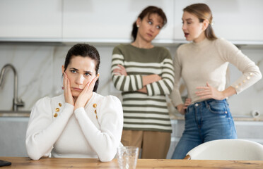 Sad middle-aged woman sitting at table and other women brawling to her standing behind in the...