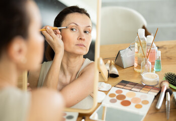 Serene woman captured in reflection of makeup mirror expertly applying eyeshadow with brush during daily beauty routine..