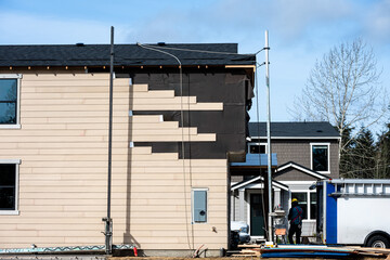 New housing construction site, now house with moisture prevention membrane and wood siding being installed with scaffolding, sunny winter day on job site, worker on ground
