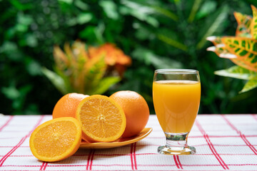 Glass cup filled with orange juice next to a yellow plate with cut oranges against a background of green plants