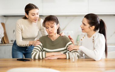 Woman is upset, sitting and grieving at table. Two female friends reassure and encourage her,...