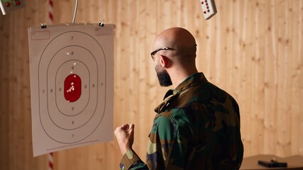 Army soldier in shooting range reloads pistol, shooting target, testing accuracy, looking at results. Military man in firing range practices with weapon, checking to see if he shot bullseye, camera B