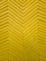 A close-up of a yellow wooden chevron pattern on an old building facade. The aged wooden planks form a symmetrical V-shaped design with visible texture and signs of wear.
