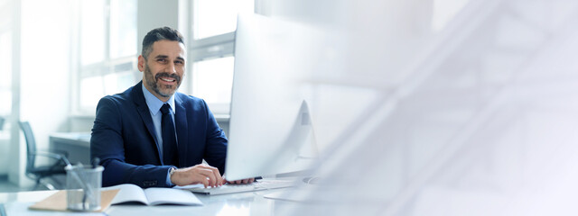 A middle aged businessman with a mature look is focused on his work at a sleek office desk. He exudes confidence while engaging with his computer. Bright natural light illuminates the space.