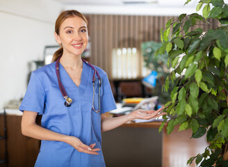 Polite smiling young woman therapist in blue uniform with stethoscope on her neck standing in medical clinic, inviting for consultation
