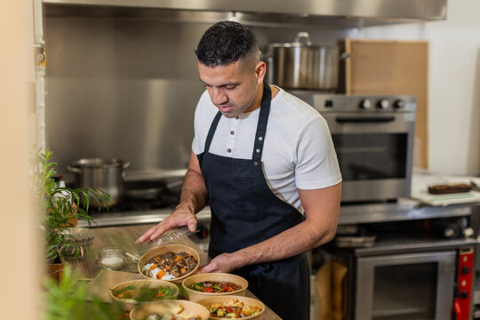 A chef wearing a black apron prepares and seals eco-friendly takeaway bowls with freshly cooked meals in a professional kitchen, ready for delivery. Horizontal, copy-space