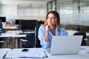 Smiling middle aged business woman using laptop looking away in office. Happy mature professional...