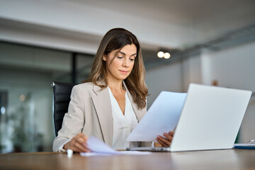 Busy serious mid aged business woman working in office with laptop reading document. Mature professional female manager lawyer attorney holding paper finance report sitting at desk in office.
