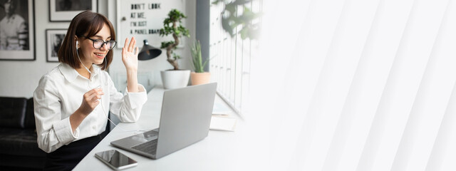 Cheerful young caucasian lady project manager having video call with client, sitting at table at cafe, waving at laptop screen, using earphones, copy space
