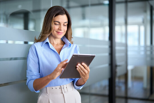 Busy middle aged businesswoman manager, older female executive leader holding pad digital tablet at work, looking at pad. Smiling mature professional business woman using tablet standing in office.