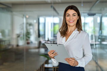 Happy 45 years old professional middle aged business woman corporate leader, happy mature female executive, lady manager standing in office holding clipboard looking at camera, portrait.