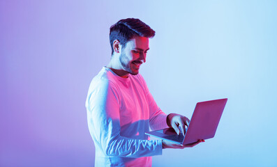 A young man stands with a laptop, engaged in his work. The background is illuminated with vibrant...