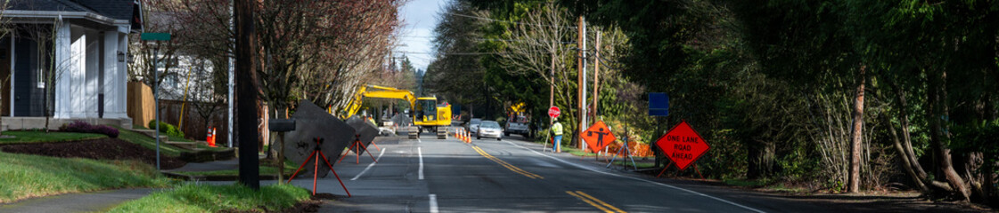Naklejka premium Public road works project, flagger managing traffic on one lane road, excavator in background for digging and trenching, winter construction project 