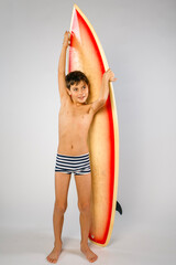 Smiling cute preteen surfer boy wearing striped swim suit standing in front of his surfboard
