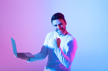 A young man in a light sweater expresses joy and triumph while using a laptop. The colorful...