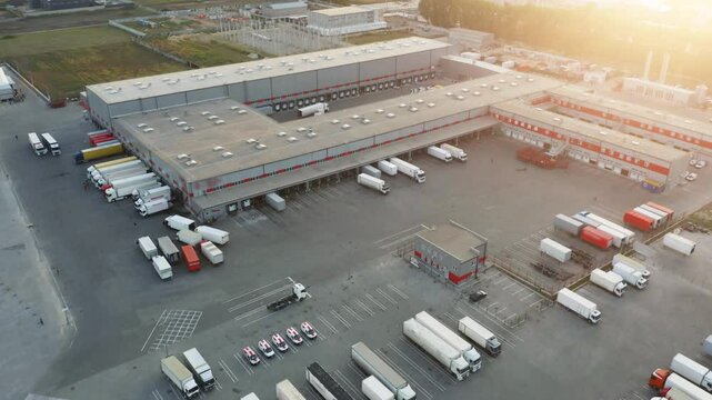 Aerial Drone View of Large Logistics Distribution Center with Warehouse Complex, Multiple Semi Trucks Loading at Shipping Docks During Golden Sunset Hour