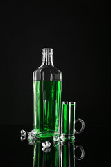 Absinthe in shot glass, bottle and ice cubes on black mirror surface