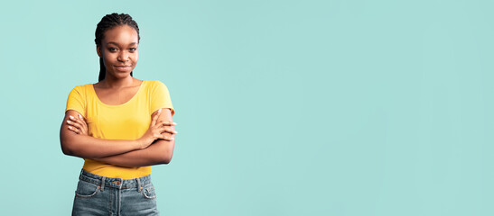 A young woman with braided hair poses confidently with crossed arms while wearing a yellow top and...