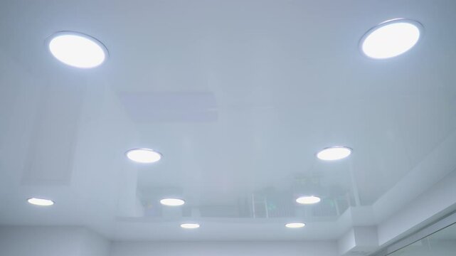 Multiple ceiling lights switch on simultaneously, illuminating a sleek modern space, reflection on glossy ceiling enhances ambient glow, creating a futuristic and minimalistic lighting effect
