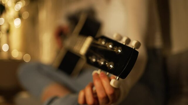 Female musician carefully tuning acoustic guitar while sitting on floor, warm lighting creating intimate atmosphere in cozy home interior with soft background illumination