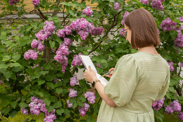 Expectant mother standing in lush garden, sketching in her art book with a pen. Surrounded by...