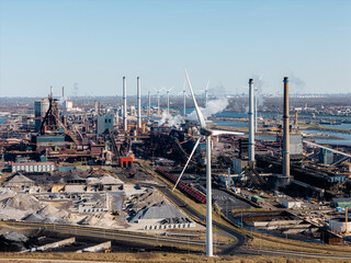 Steel production site in IJmuiden, Netherlands, featuring smokestacks, raw materials, and wind turbines. The aerial perspective highlights the tension between heavy industry and renewable energy.