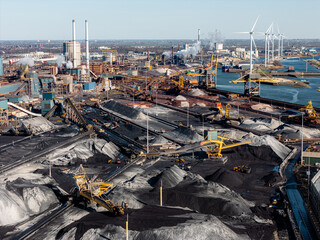 Steel industry in IJmuiden, Netherlands, showing coal storage, conveyors, and emissions. Wind turbines stand in contrast to fossil fuel-based production and industrial pollution. Aerial view.