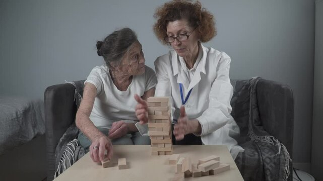 Senior woman practice skills build wooden blocks, building tower and trying not to let it fall, Jenga game. Old patient pull out block, place on top, support dotor during therapy dementia in house.