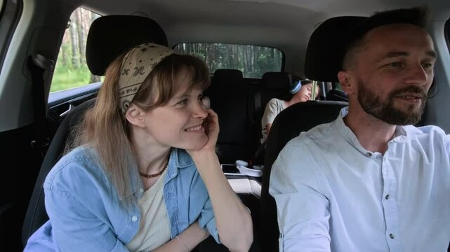 Man driving car, smiling and chatting with his wife in passenger seat while children sleeping in backseat on road trip through forest