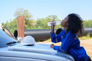 A black female engineer takes a break while working in the hot sun on an industrial pipe work site.