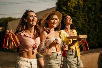 Cheerful women carrying pizza and drinks laughing together at a party